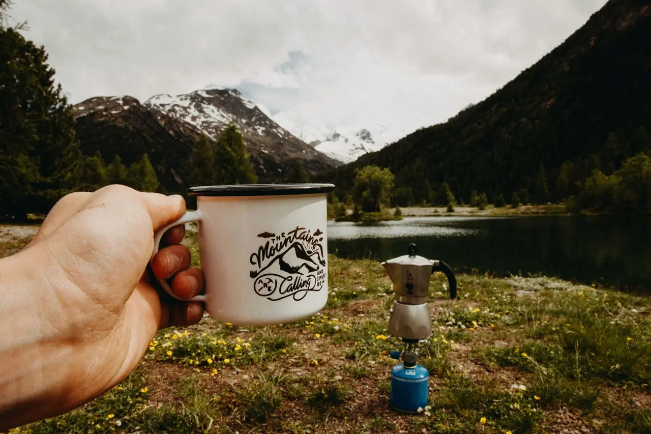 A camp mug held up in front of a mountain lake
