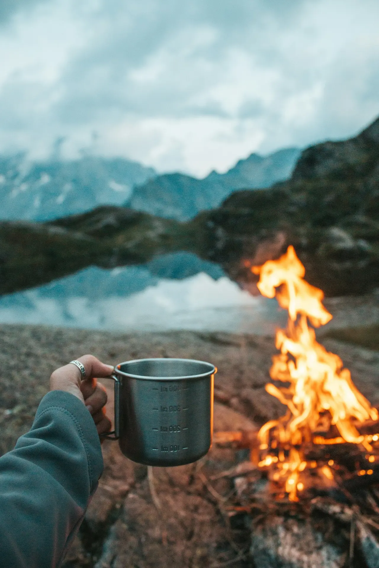 A hand holding a camp mug beside a campfire and a lake